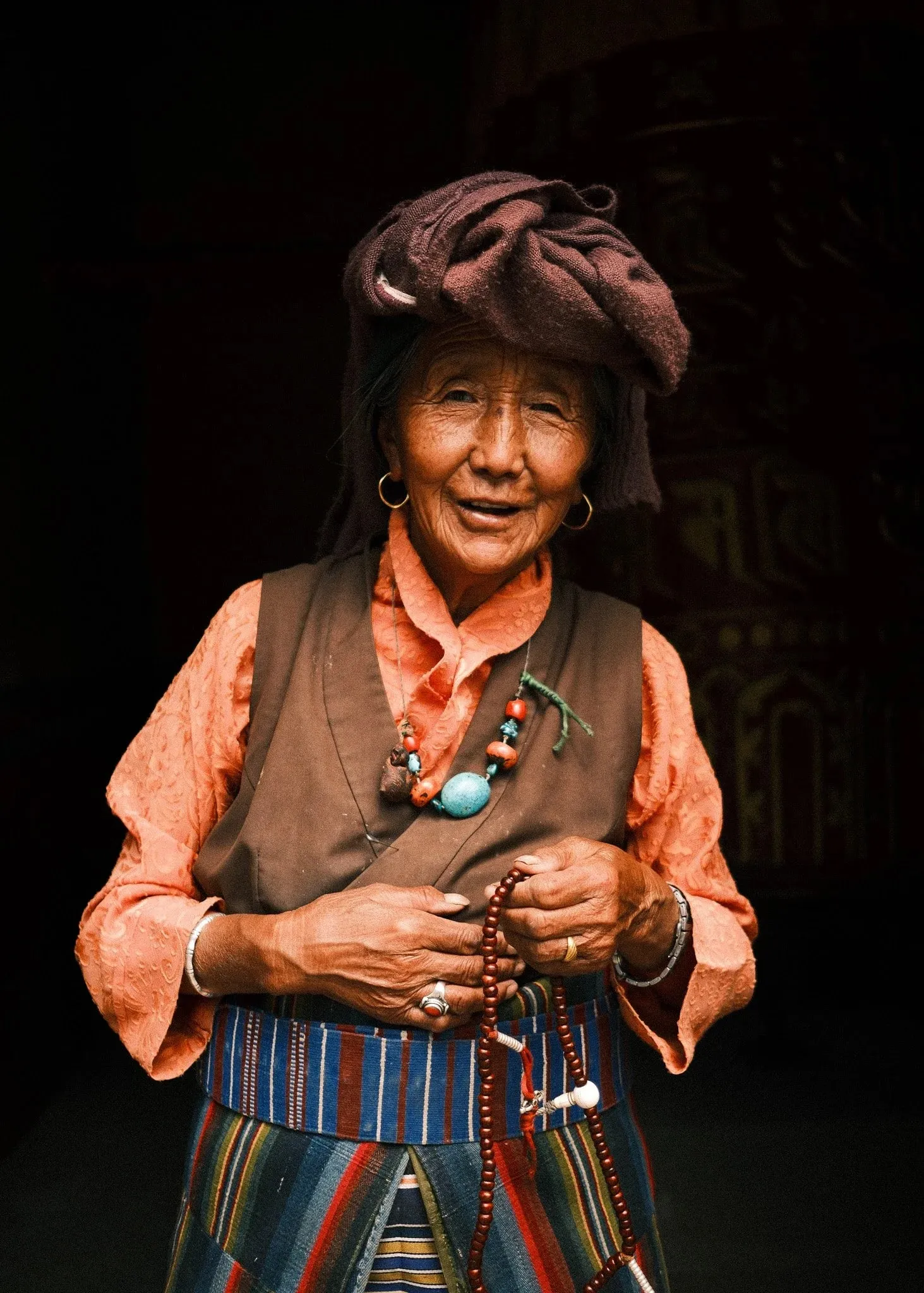 Older woman wearing a dark headwrap and turquoise necklace, holding prayer beads in Tsarang, Upper Mustang, Nepal.