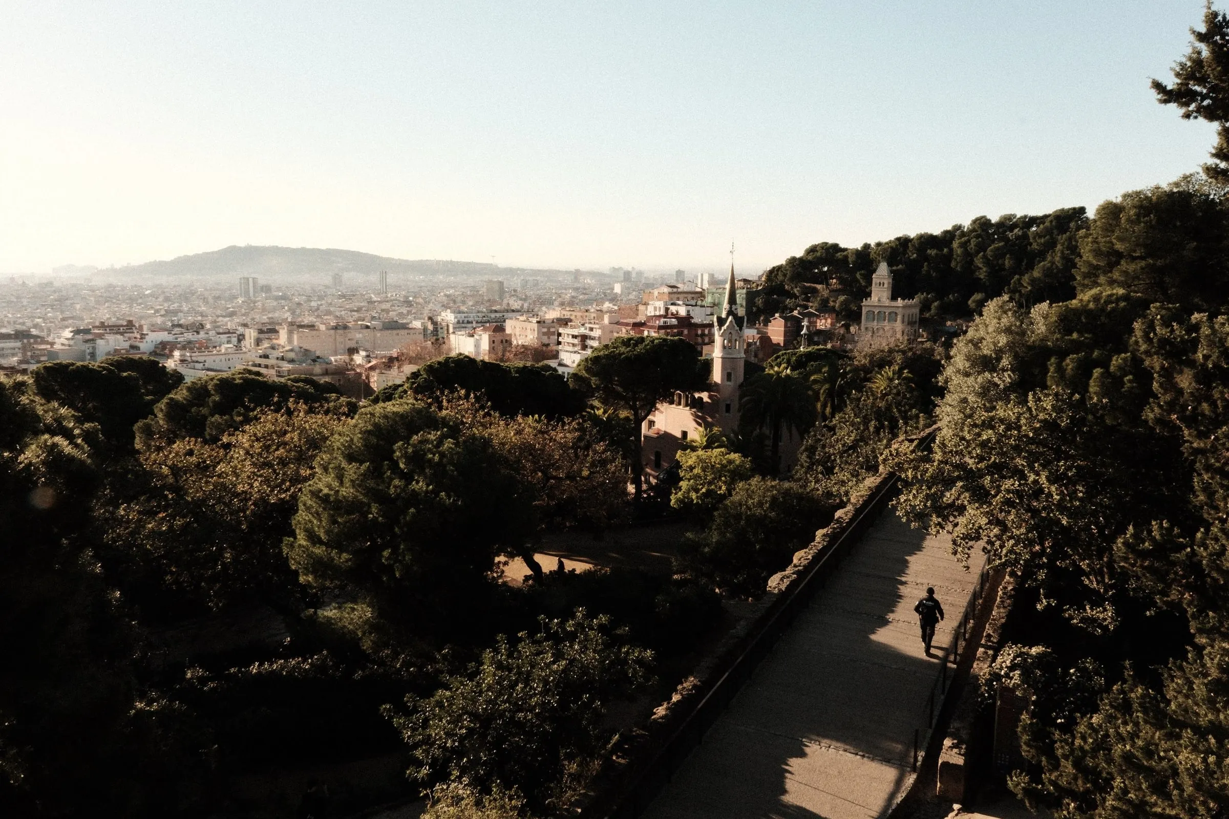 A person walks along a sunlit path through the trees of Park Güell, with the city beyond