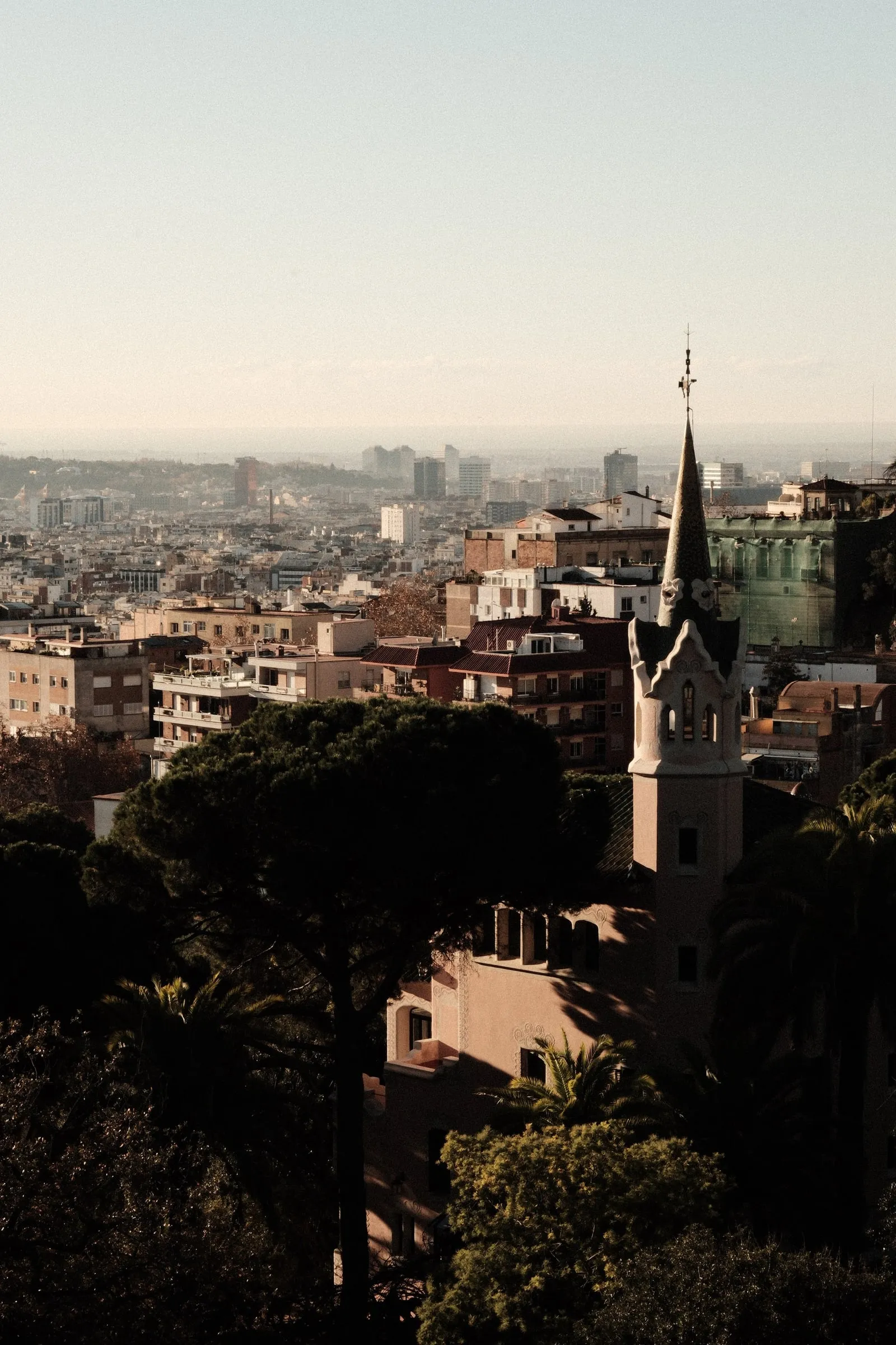 A pointed tower rising above trees, with the city stretching towards the sea, seen from Park Güell
