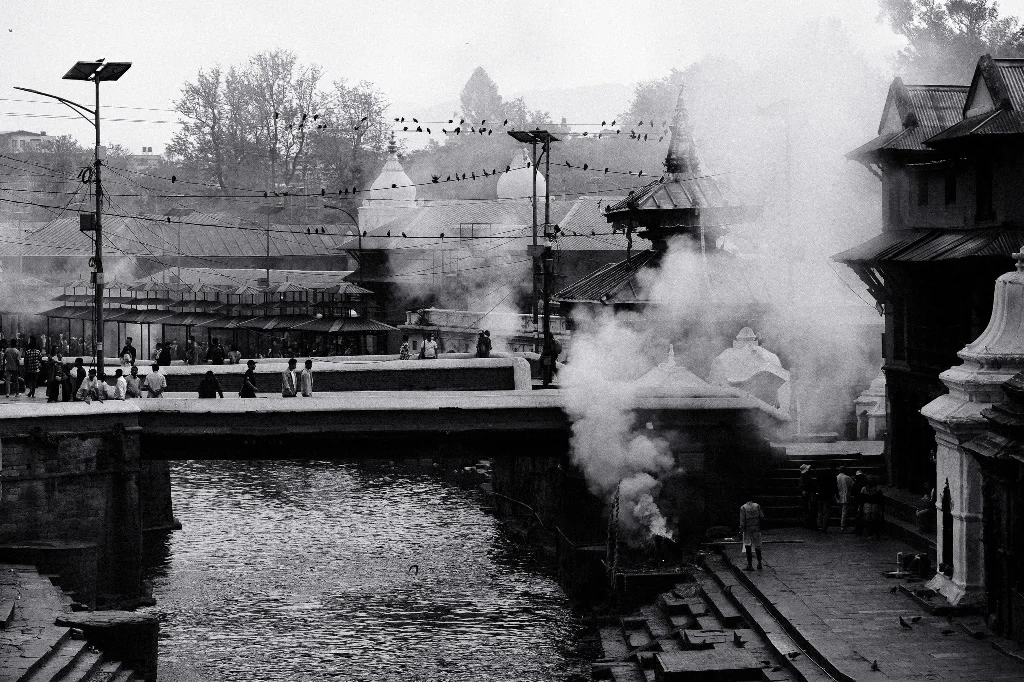 Black and white photo of smoke rising from the cremation ghats beside the river at Pashupatinath