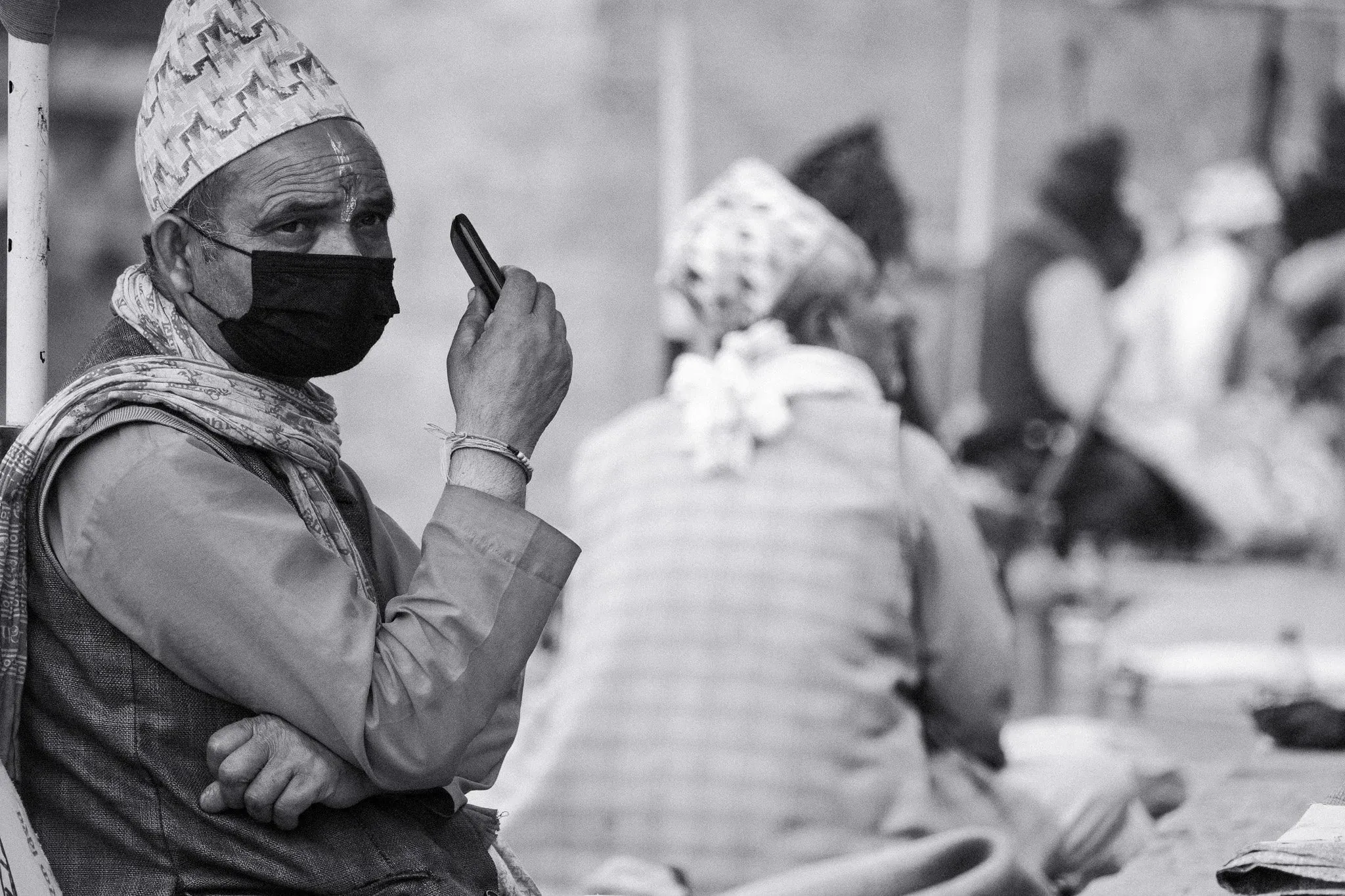 Black and white photo of a man in a topi and face mask sitting at Pashupatinath