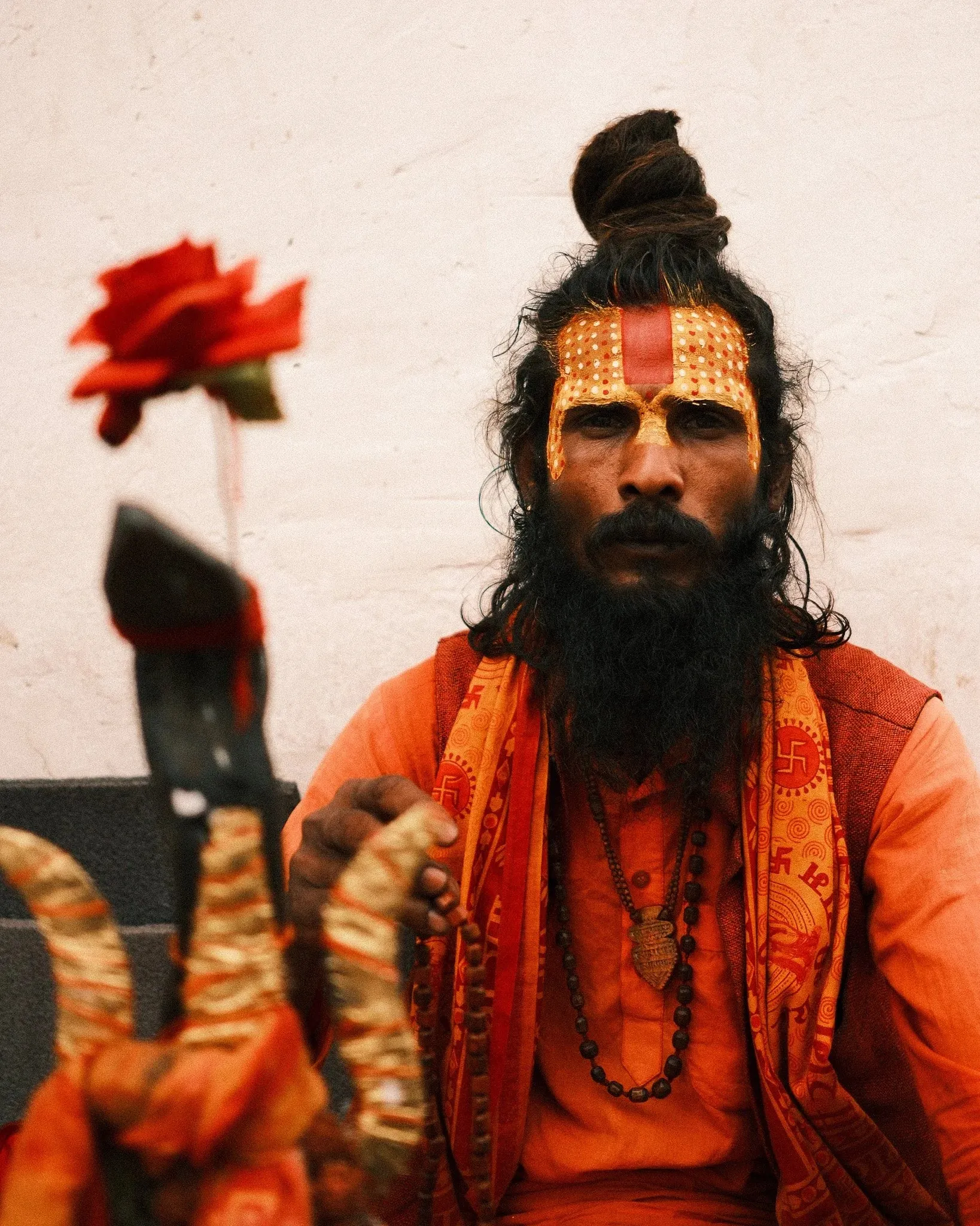 A sadhu with a painted face and orange robes holding a red flower at Pashupatinath