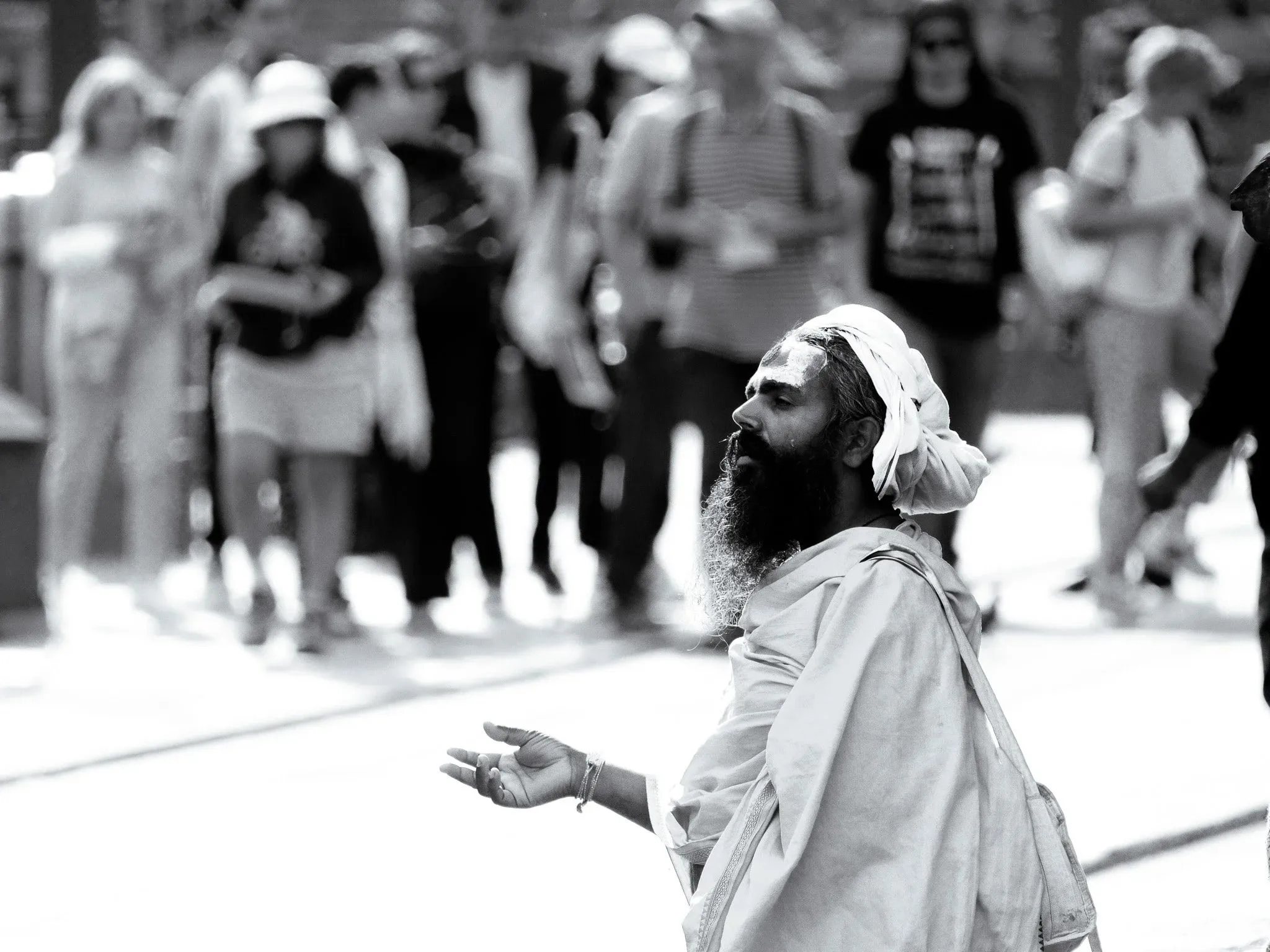 Black and white photo of a sadhu holding out his hand as a group of tourists walks past