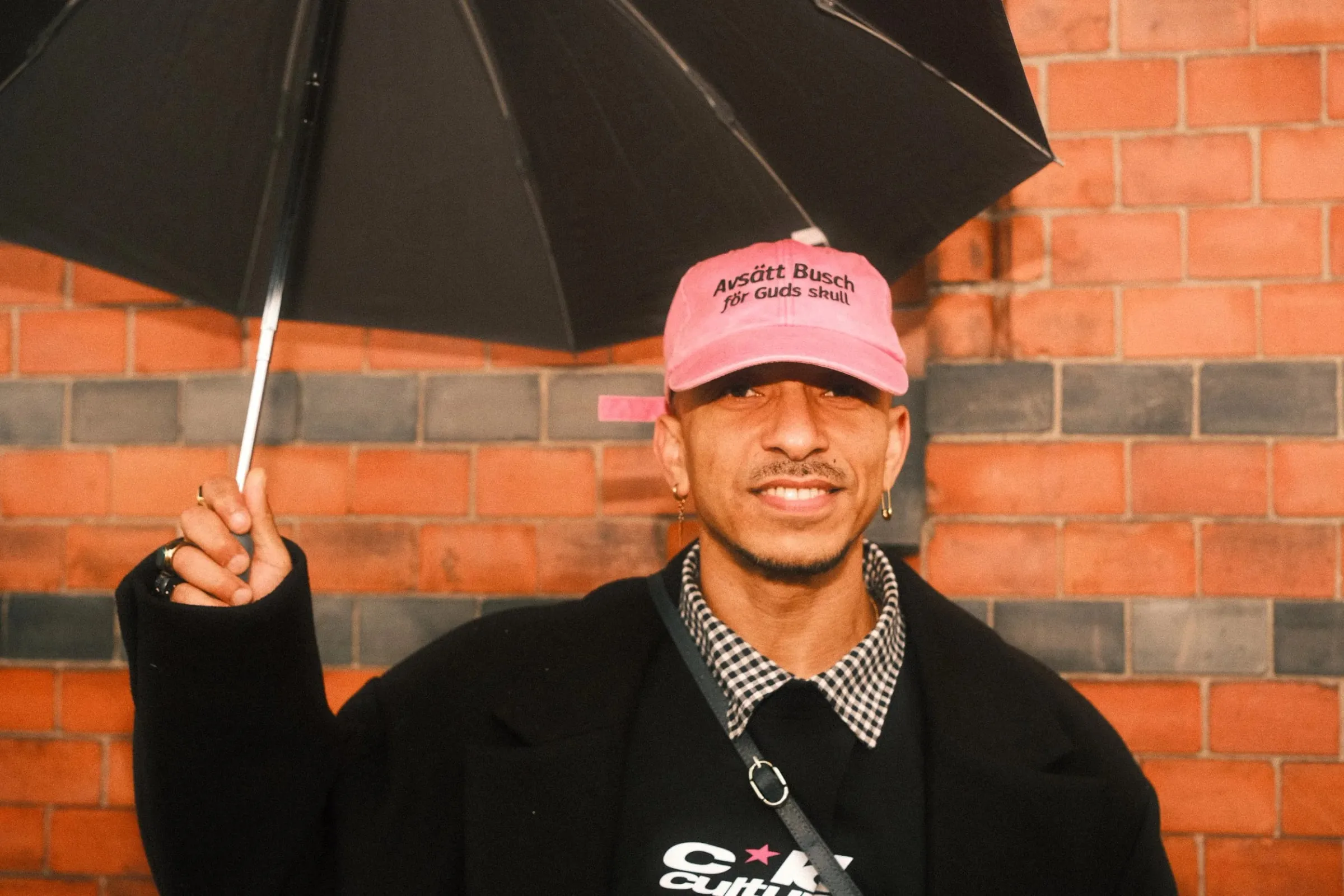 A smiling man in a pink cap holding an umbrella in front of a brick wall