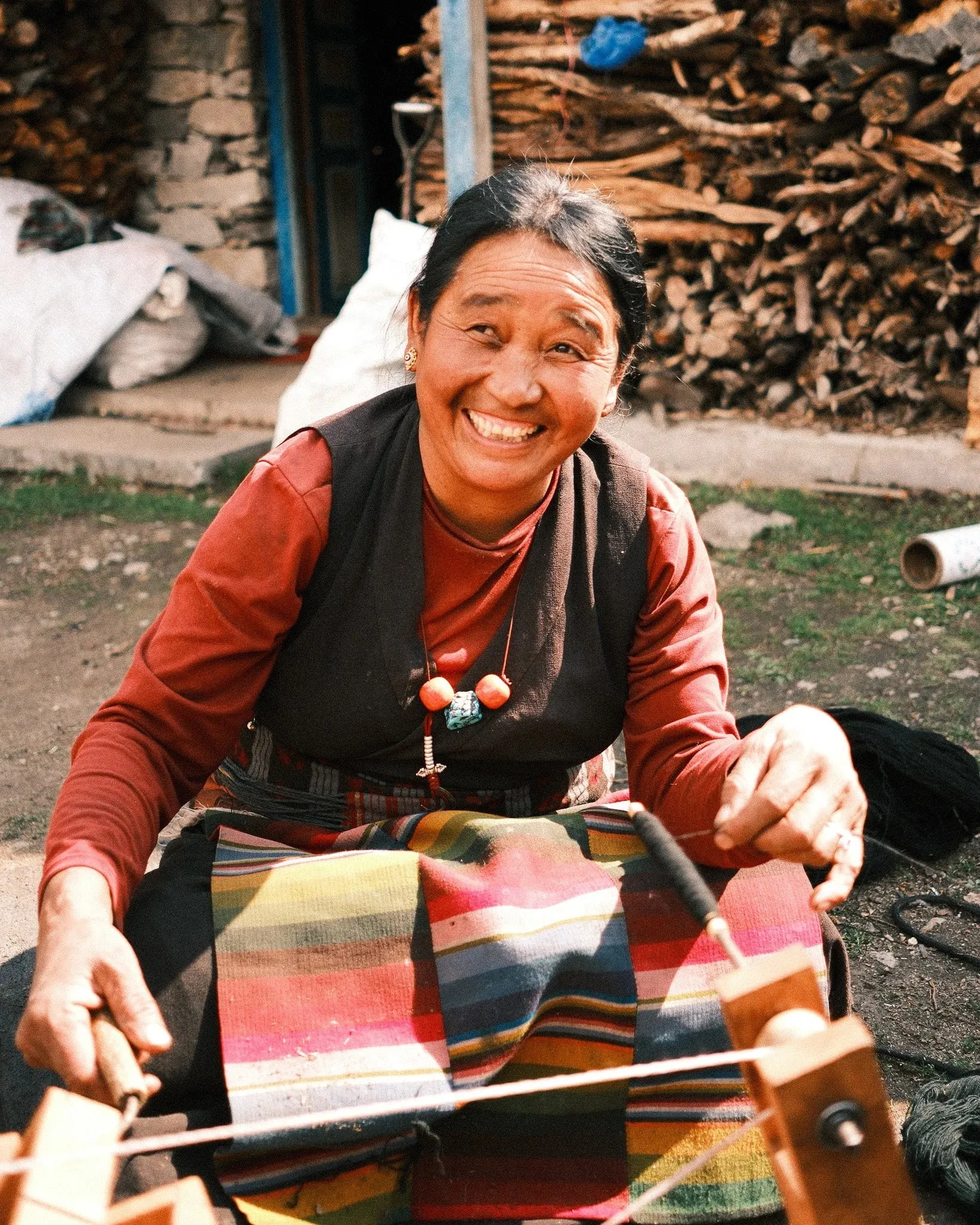 A smiling woman weaving striped cloth on a small loom outside her home