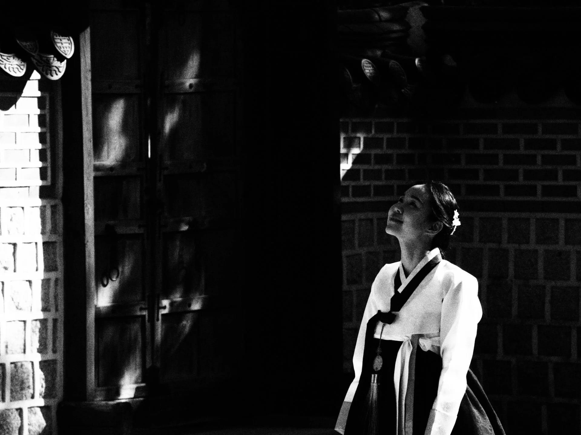Woman in hanbok standing in a patch of light beside a dark doorway