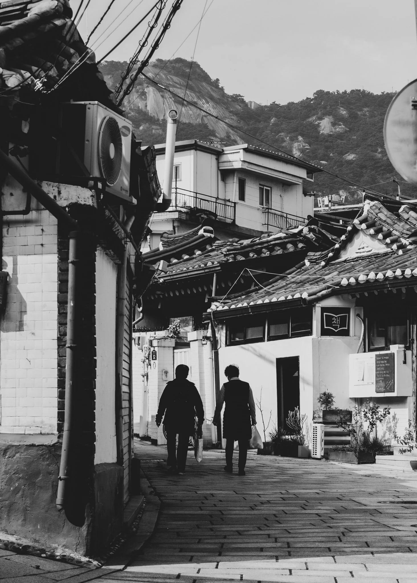 Two people walking up a narrow street of old houses toward a mountain