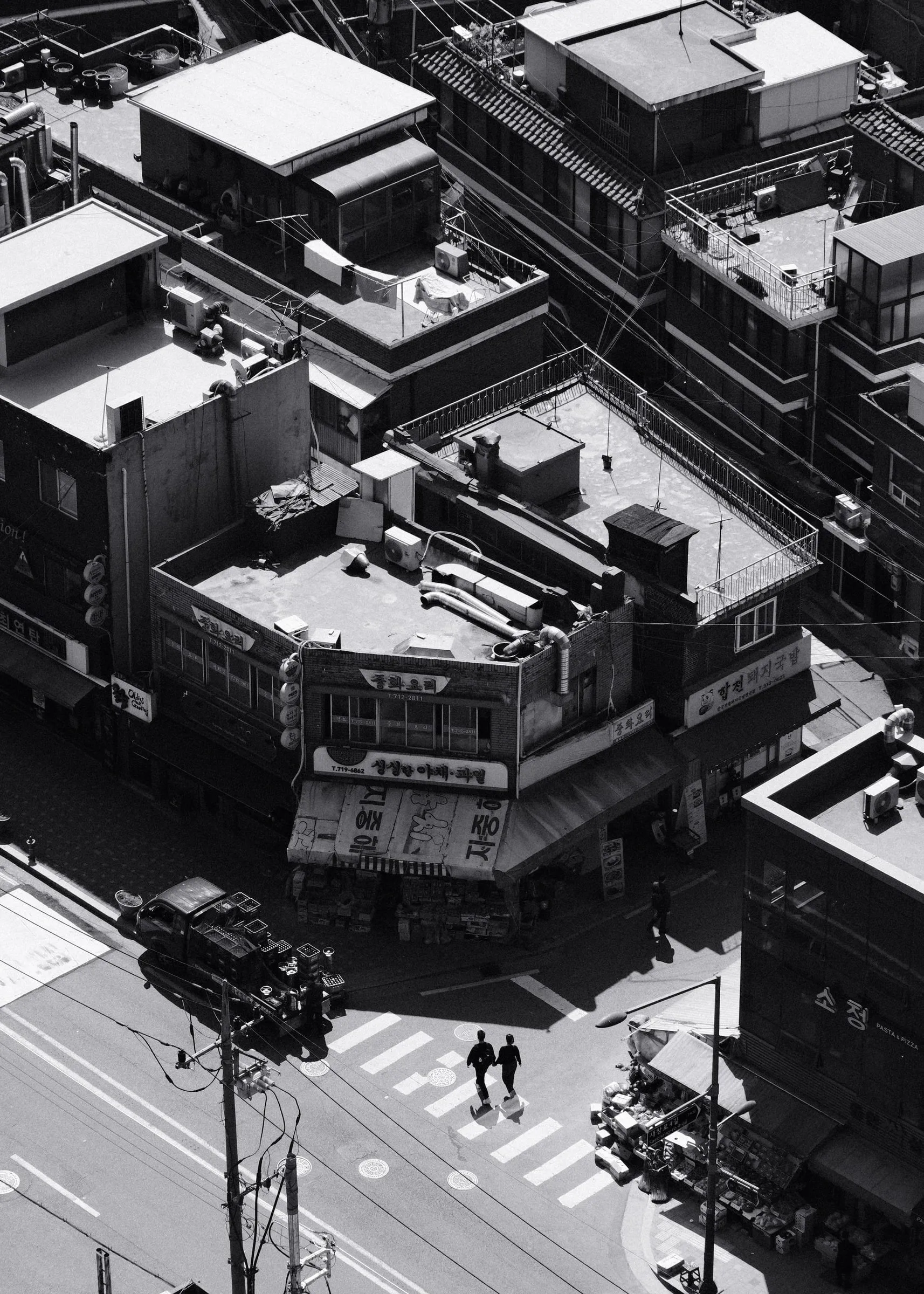 Black and white view from above of two people crossing a street between low rooftops in Seoul