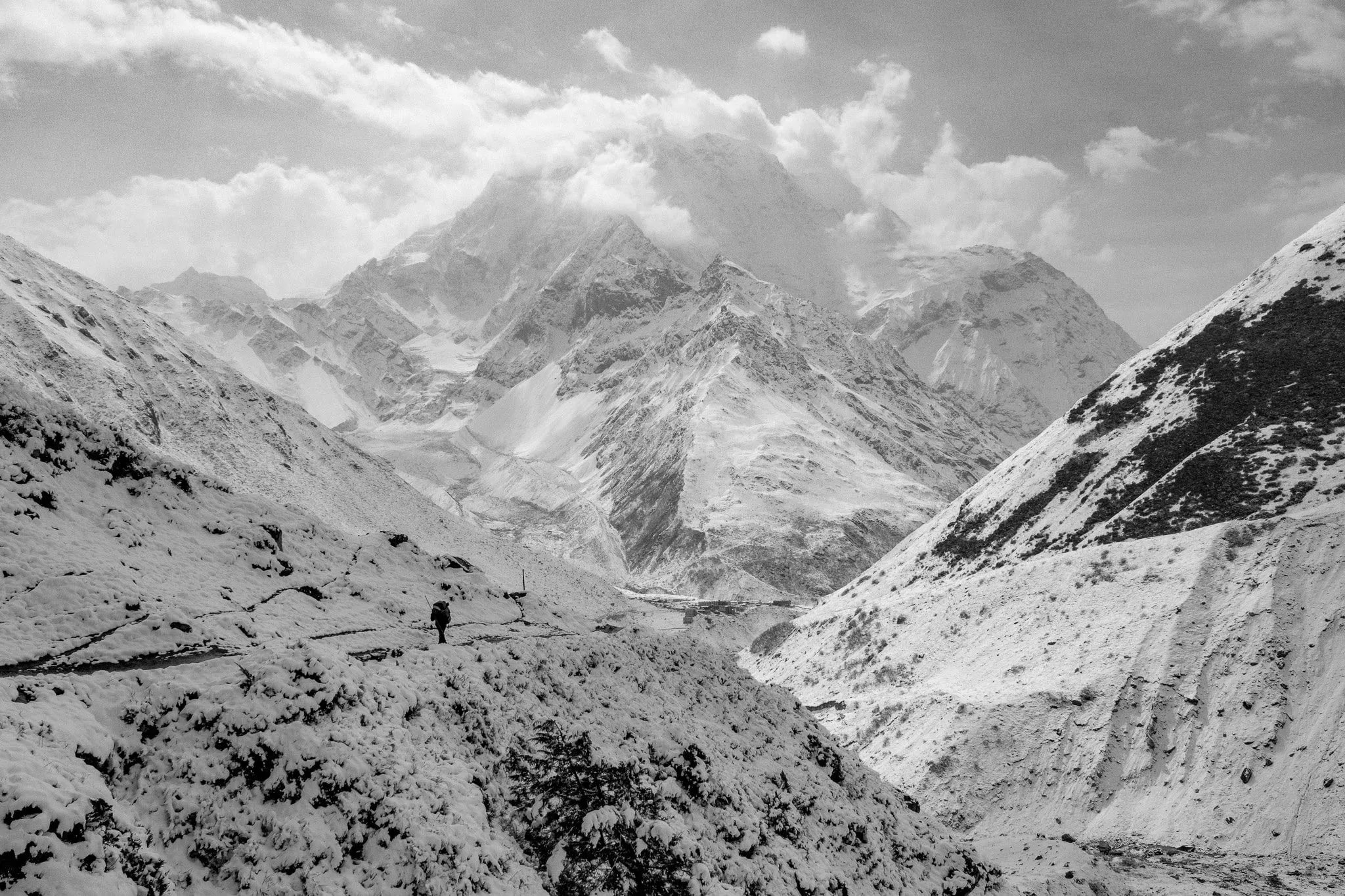 Black and white photo of a lone trekker crossing a snowy valley beneath the peaks