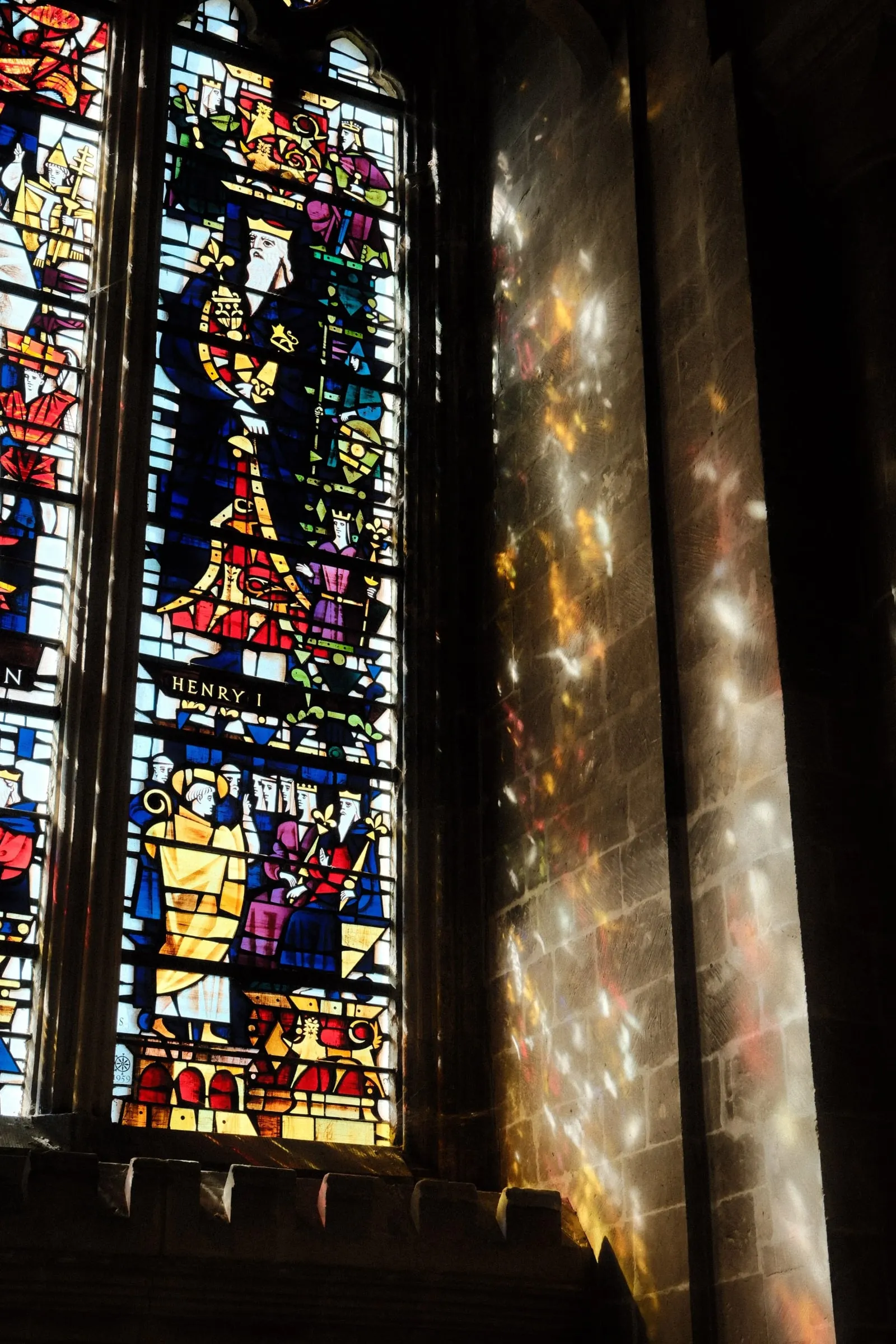 A stained glass window casting patches of coloured light onto the stone wall beside it