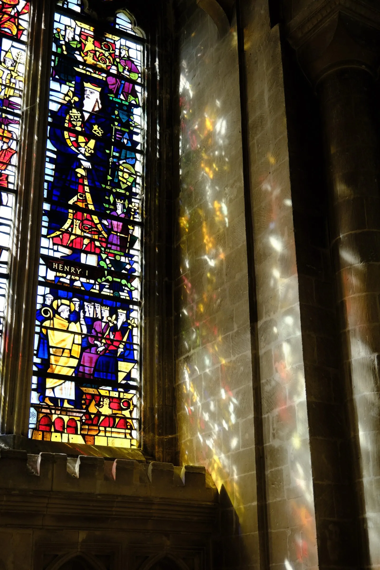 A stained glass window casting patches of coloured light onto the stone wall beside it