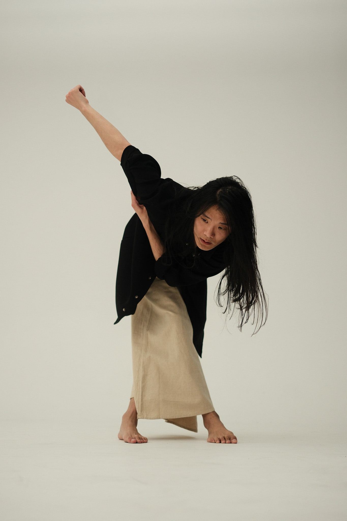 Dancer in a black jacket and cream trousers kicking one leg high against a pale backdrop