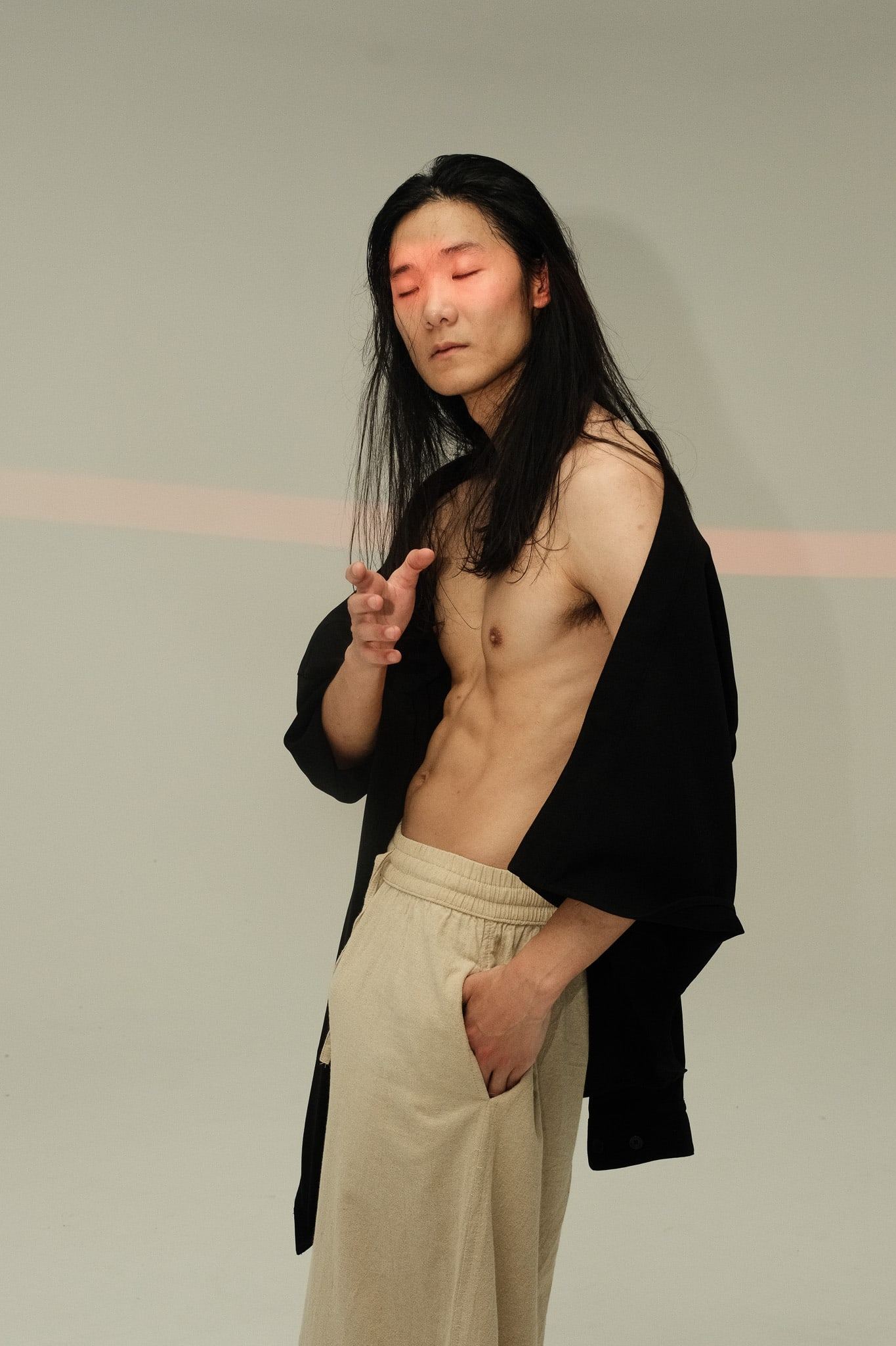 Shirtless model in an open black jacket and cream trousers, eyes closed, one hand raised