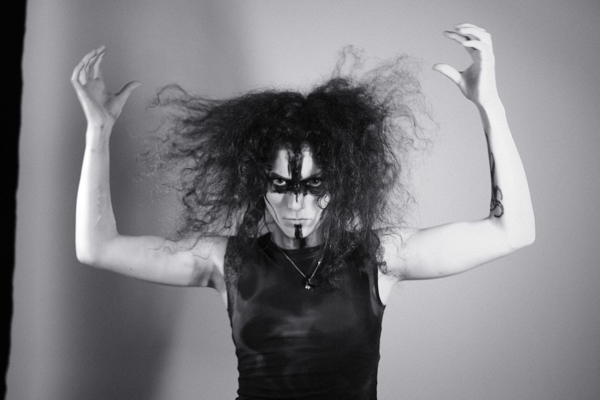 Black-and-white portrait of a model with face paint and voluminous hair, arms raised above the head