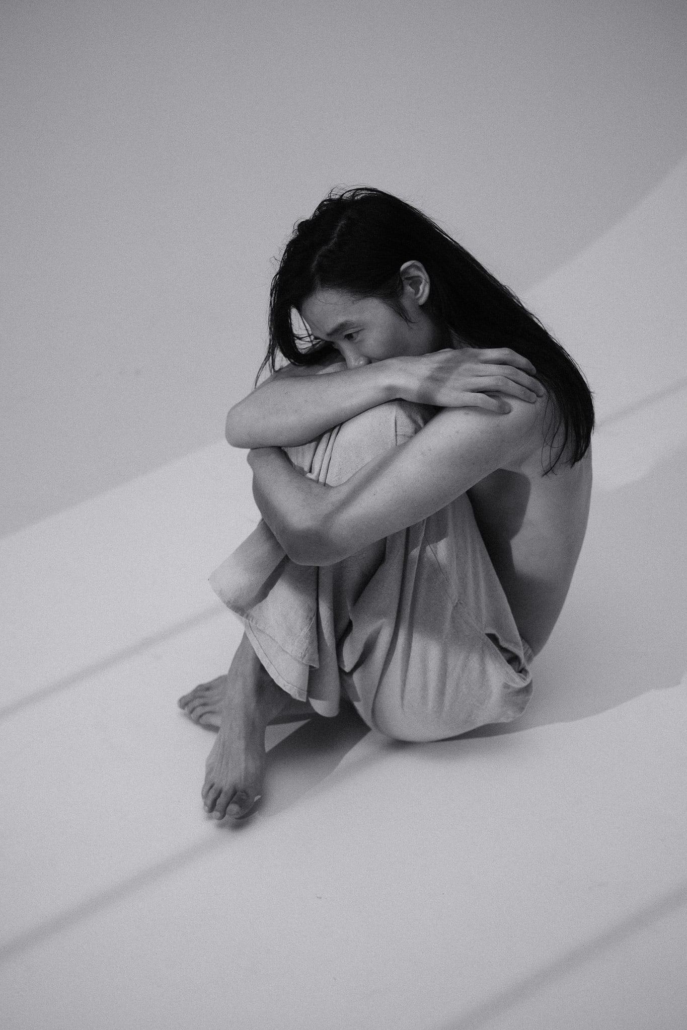 Black-and-white portrait of a person hugging their knees on the studio floor