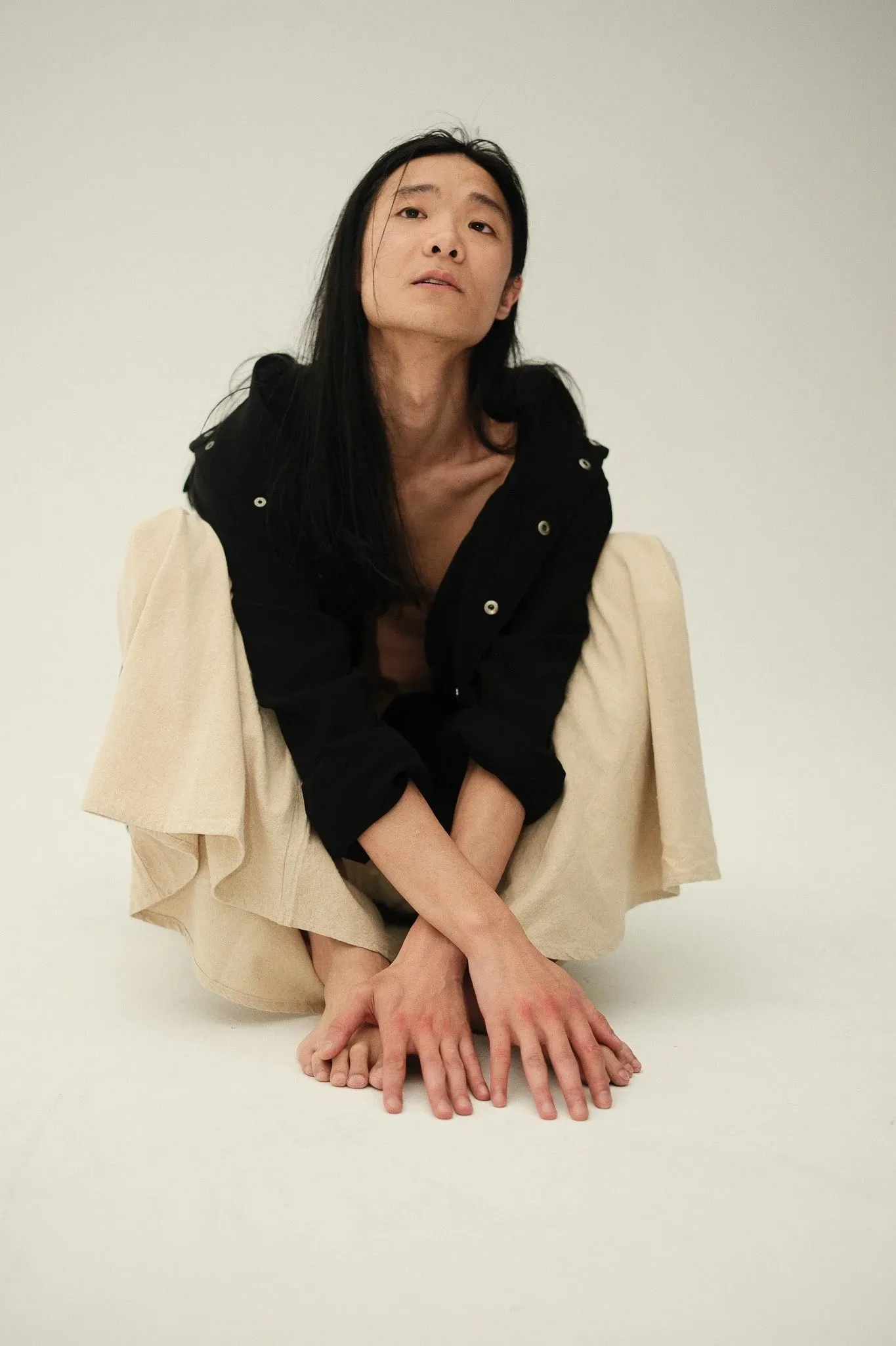A model with long dark hair crouching on the floor in a black shirt and cream trousers