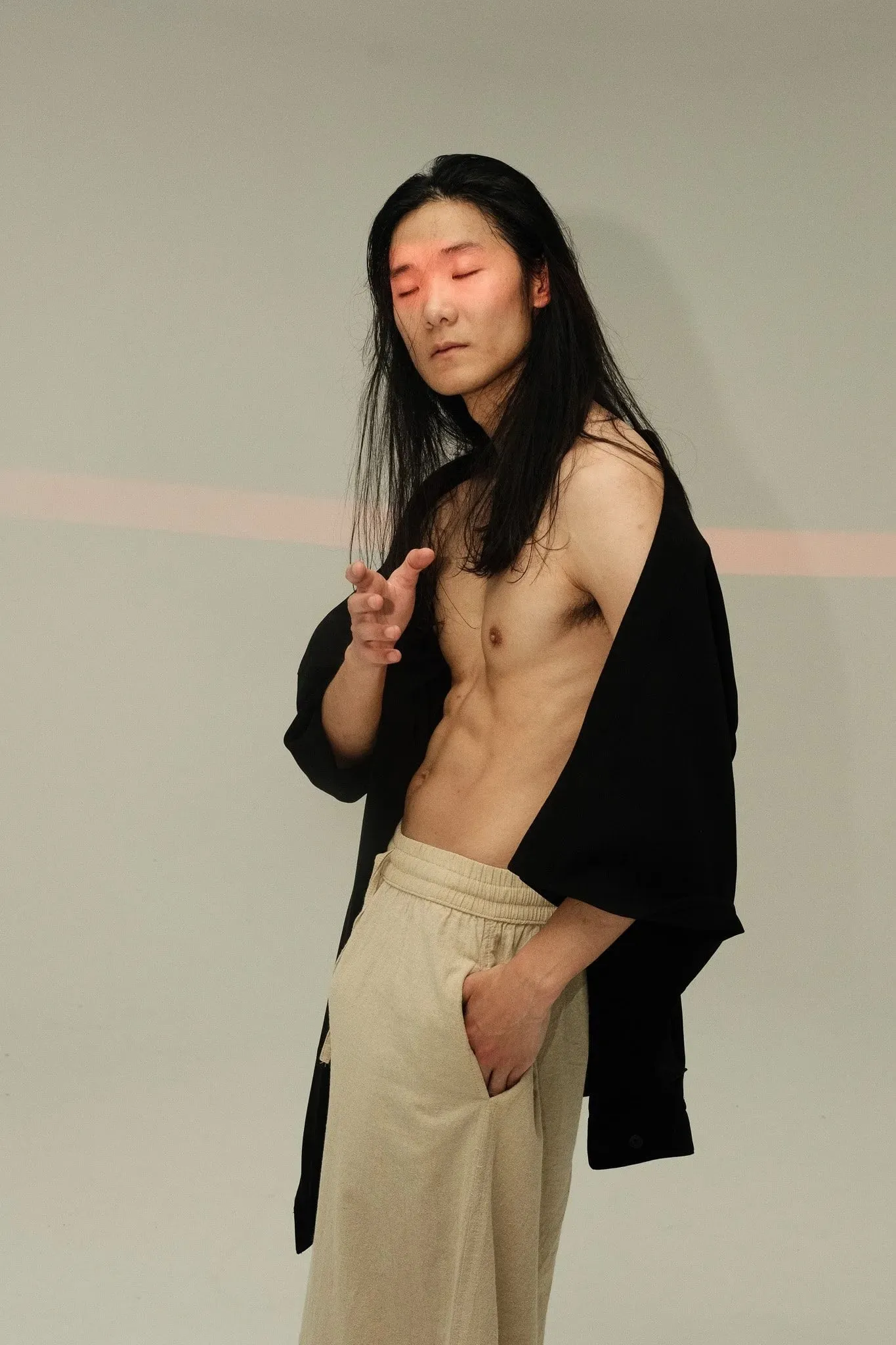 A model in an open black shirt and cream trousers standing against a pale wall