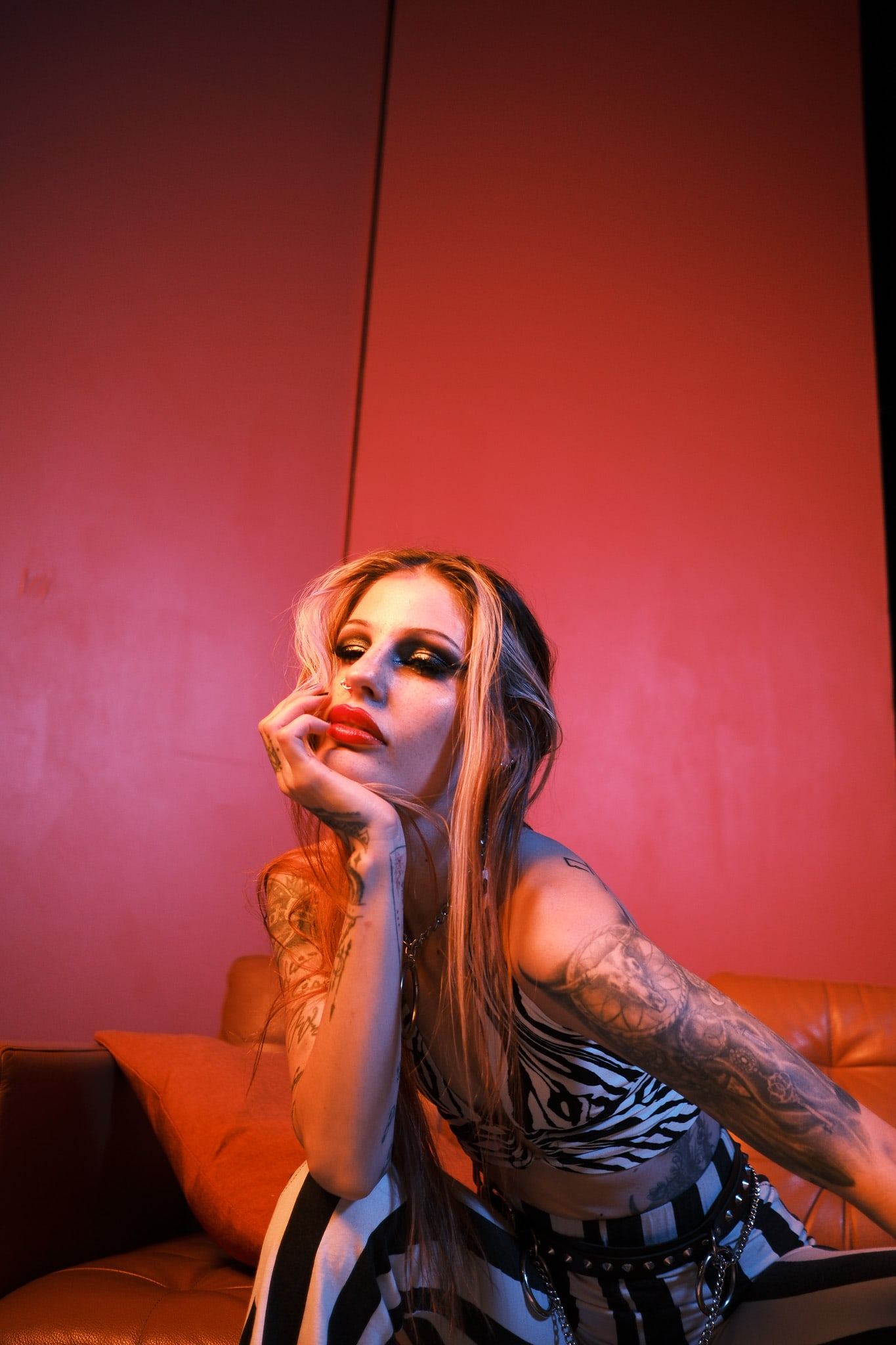 Woman with tattooed arms leaning on an orange sofa under red light, one hand at her chin