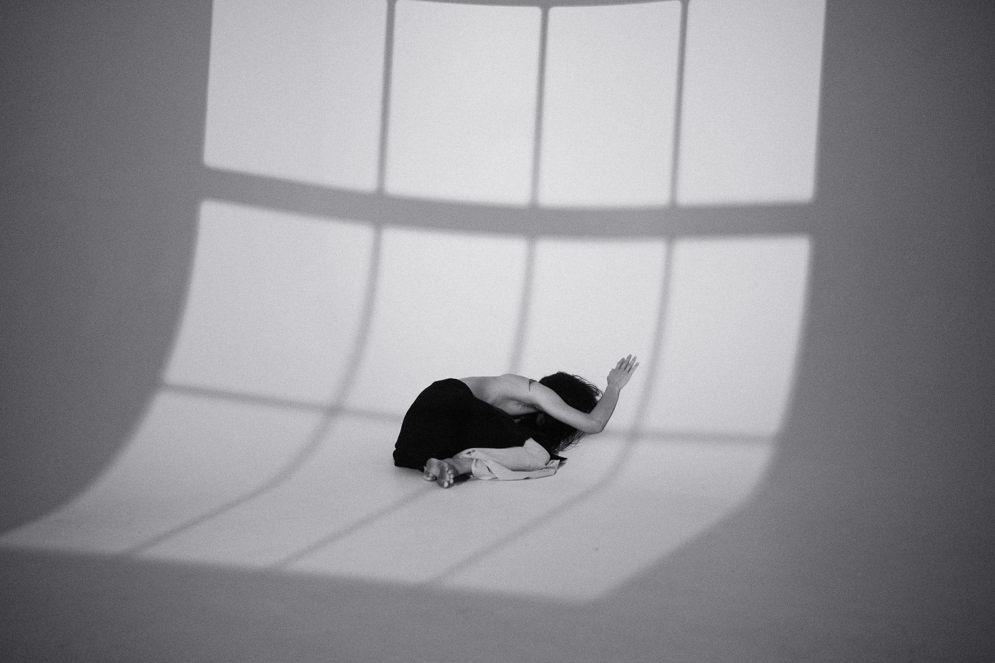 Figure curled up on a white studio cove crossed by the shadow of a window frame