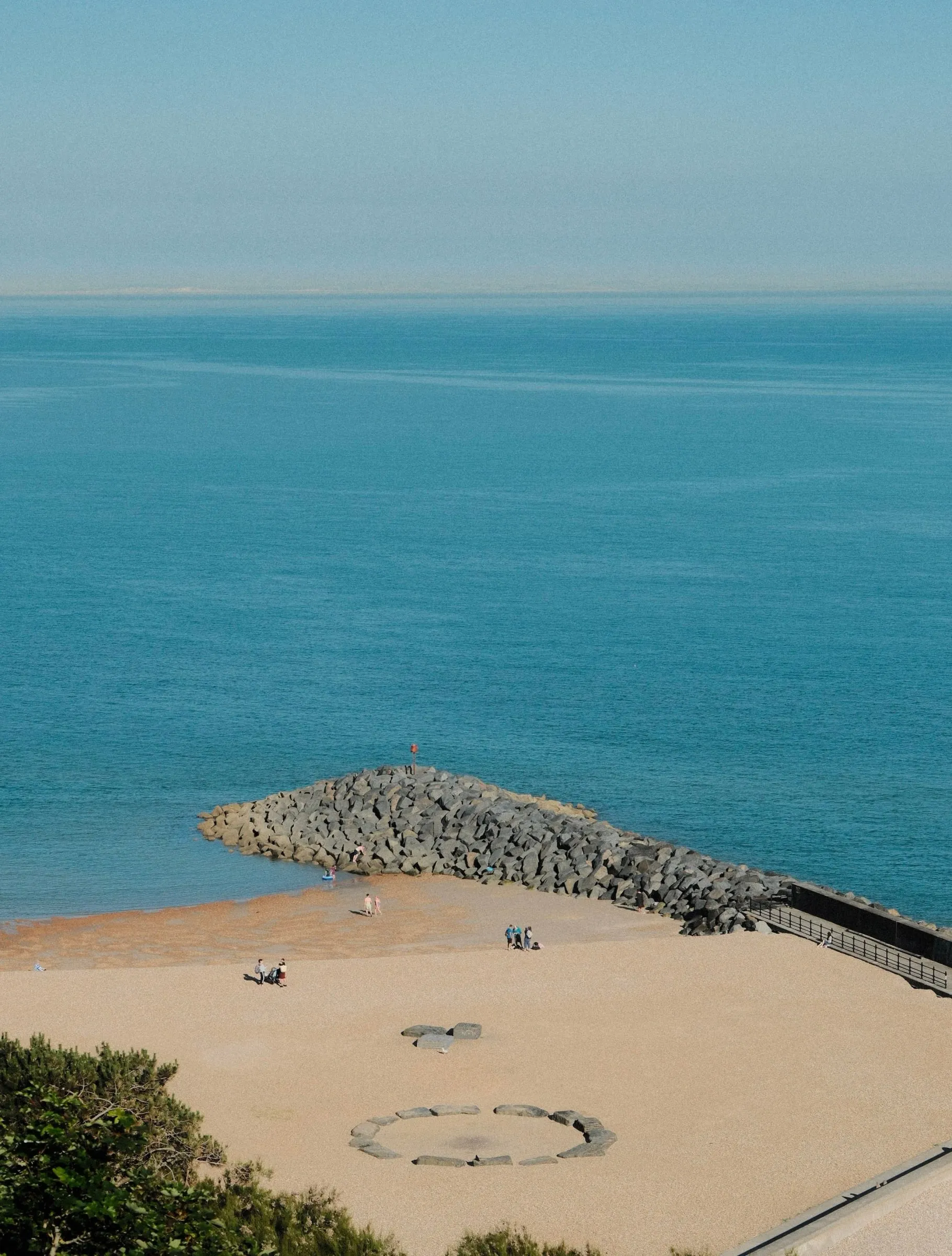 A sandy beach and rock groyne seen from above, with a calm blue sea