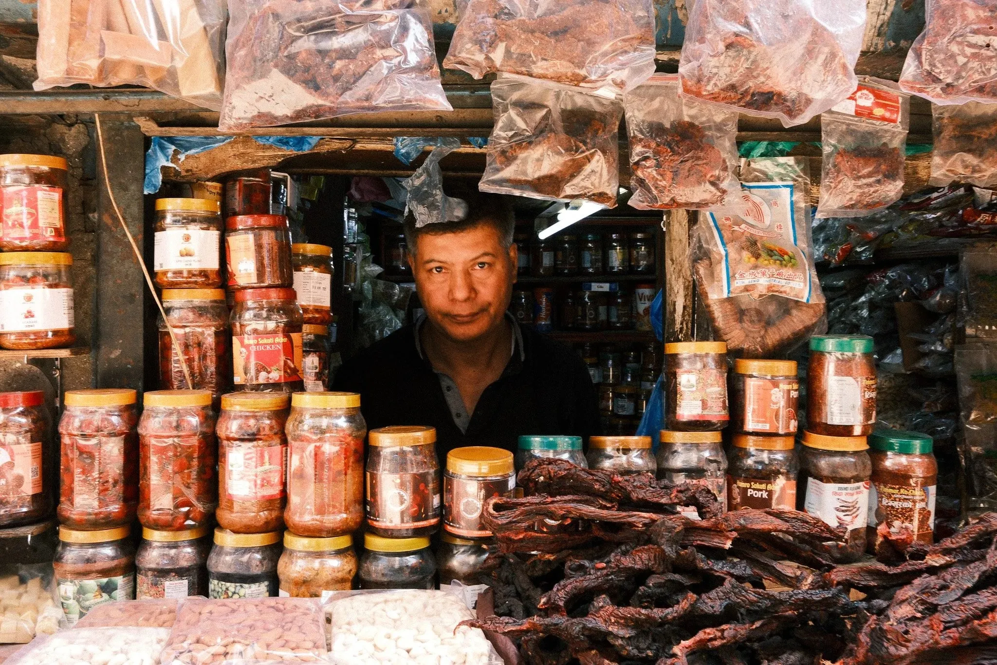 A shopkeeper looking out from behind jars of pickles and hanging bags of spices