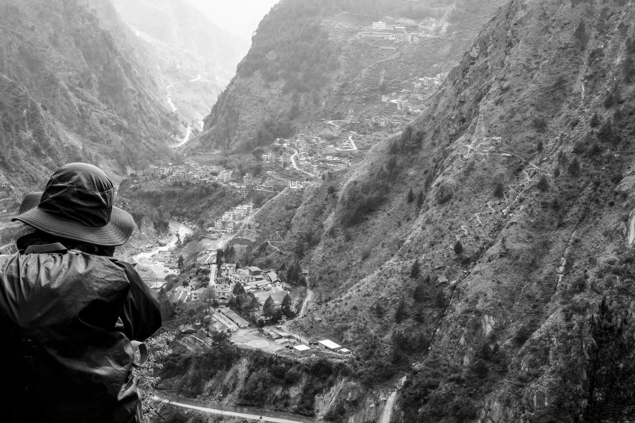 Black and white photo of a person in a hat looking down into a steep valley with a village below