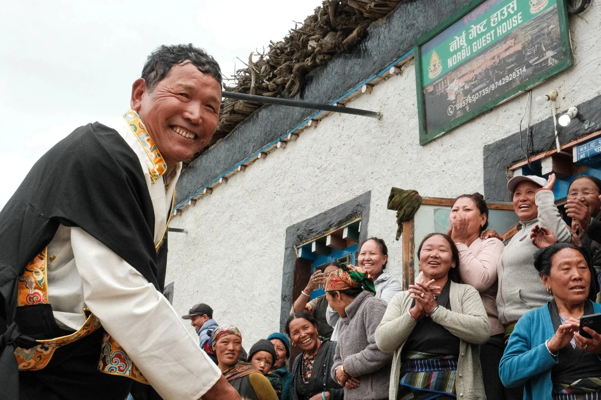 A smiling man in traditional dress at the front of a crowd of laughing villagers in Tsarang