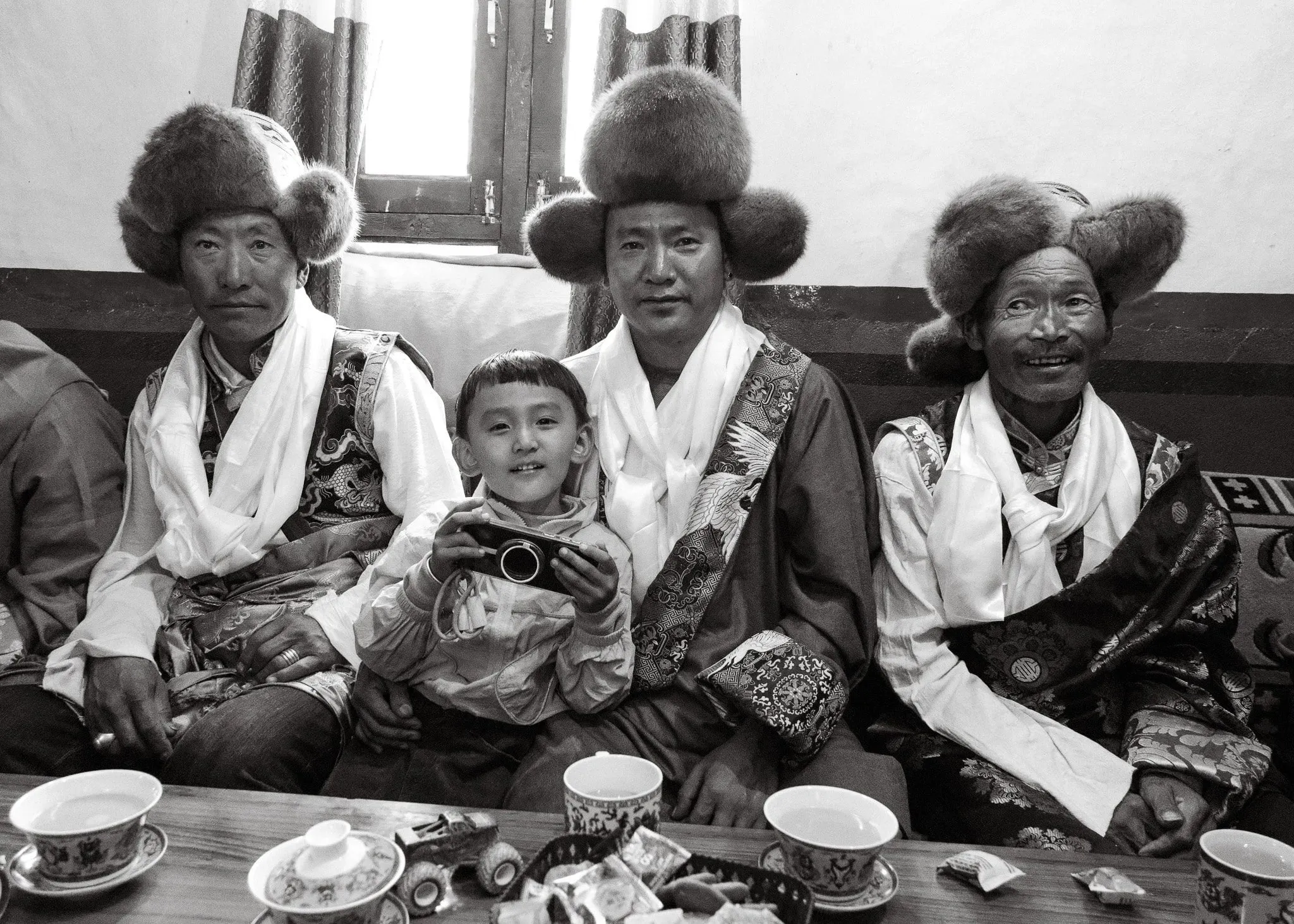 Black and white photo of men in fur hats and white scarves sitting with a young boy in Tsarang