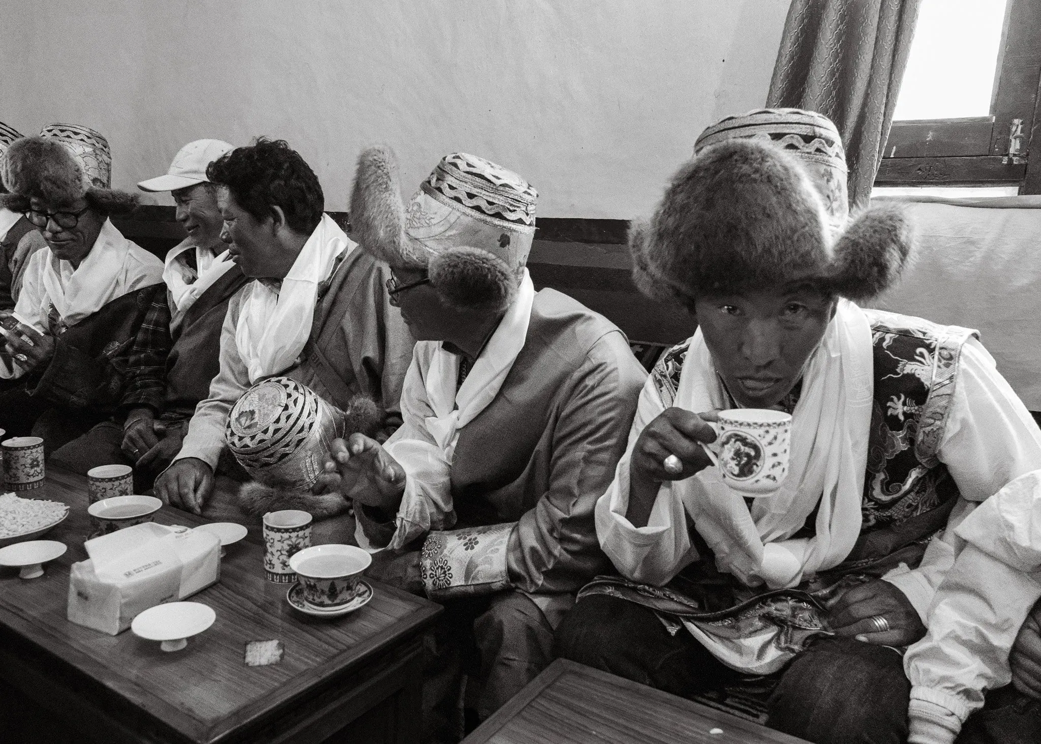 Black and white photo of men in fur hats and white scarves drinking tea at a table in Tsarang