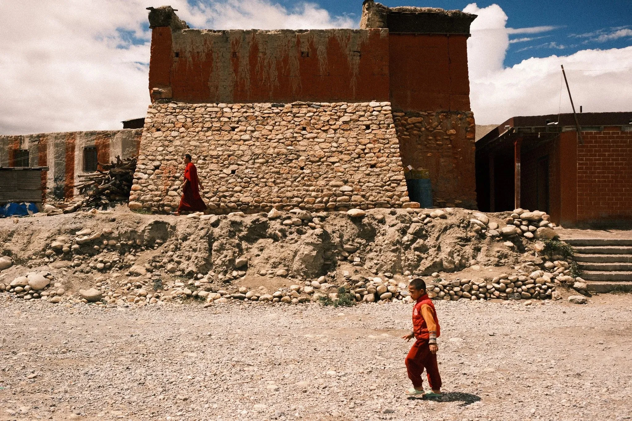 A young monk walking across open ground below an old stone building in Tsarang