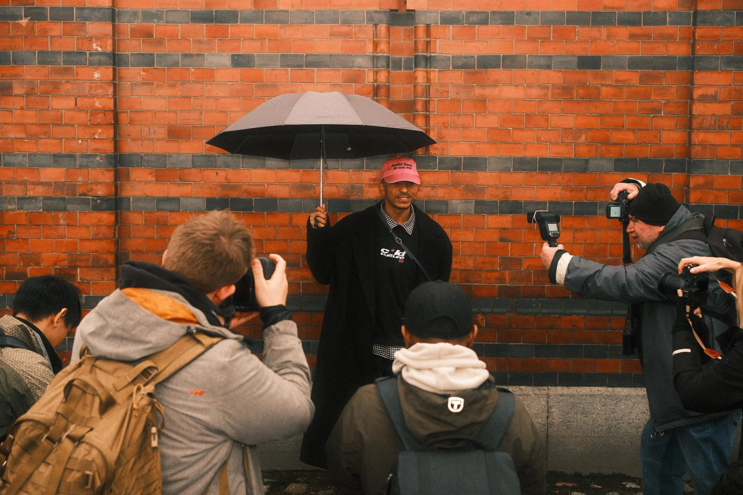 Photographers gathering round a man in a pink cap holding an umbrella against a brick wall